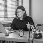 Black and white photo of a woman working at a desk with a laptop and vintage telephone.